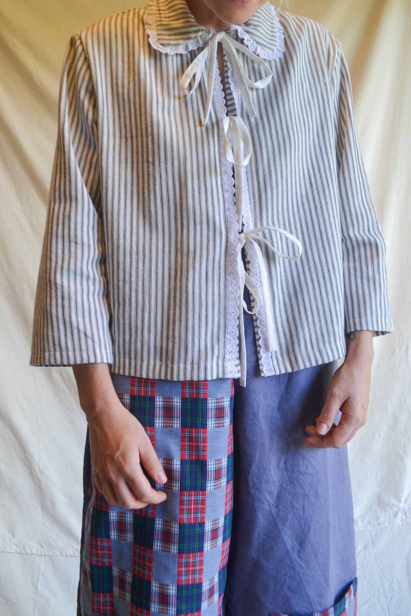 Person wearing a striped shirt with a lace collar and plaid pants against a plain background